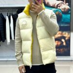 Mon蒙口（鹅绒）、2025秋冬新品 | MONCLE羽绒马甲。

米白色立领设计搭配顺滑拉链系统，正面采用100%锦纶防风面料，背面运用亮黄色轻量材质形成撞色美学。填充90%鹅绒与10%羽毛的黄金配比，绒朵蓬松度达600+FP，充绒量精准适配5℃至-5℃环境。左胸点缀经典立体刺绣徽章，两侧斜插口袋以隐藏式拉链保持流畅线条。

双面不同配色实现一衣多穿场景：米白面可搭配同色系针织衫演绎都市极简风，亮黄面翻转后瞬间提升视觉活力。立体剪裁确保活动自由度，台湾盟可睐有限公司精工制造，通过RN116347质量追溯体系。从防泼水锦纶到鹅绒填充，诠释蒙口将高山机能基因融入日常穿着的设计哲学。1-5码（模特173，143斤上身2码） - Image 2